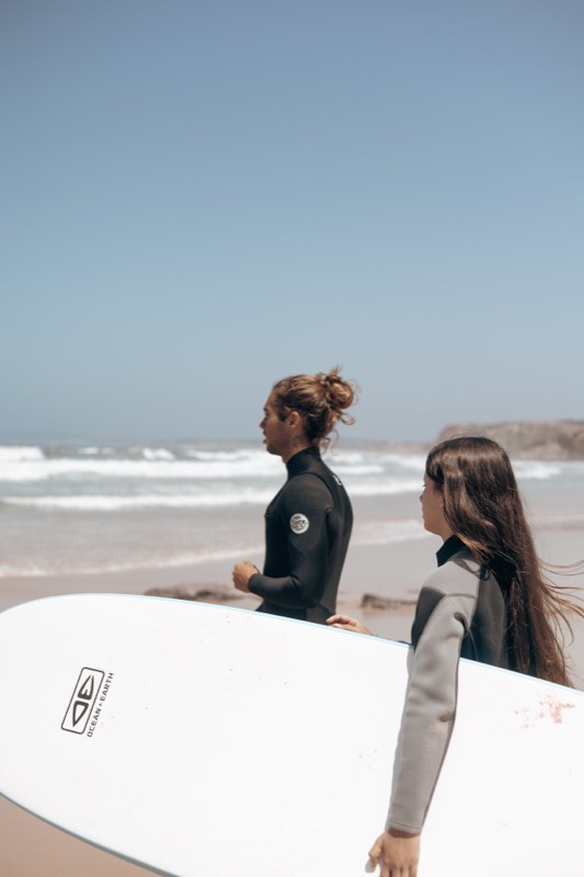 Two surfers in wetsuits walking down the beach with their boards, the cliff behind them.