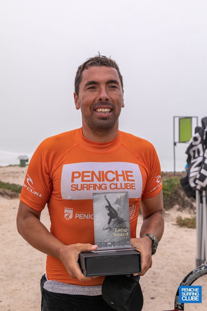 Bruno Grandela in a Peniche Surfing Clube jersey holding a glass longboard trophy on the beach.