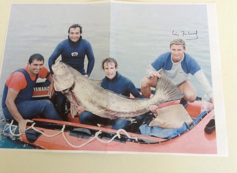 Four men in wetsuits on an orange inflatable boat holding a corvina almost as long as they are tall.