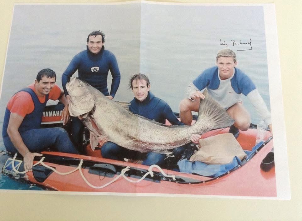 Four men in wetsuits on an orange inflatable boat holding a corvina almost as long as they are tall.