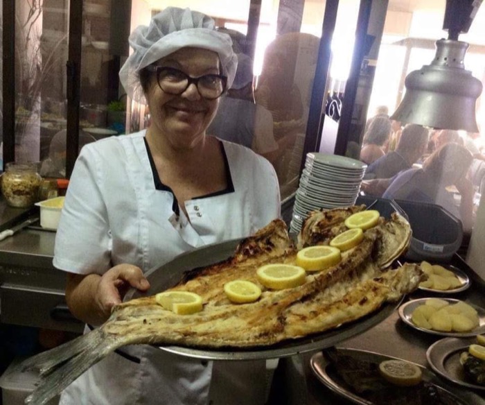 My grandmother in chef whites and a hairnet, smiling, carrying a large platter of grilled fish with lemon.