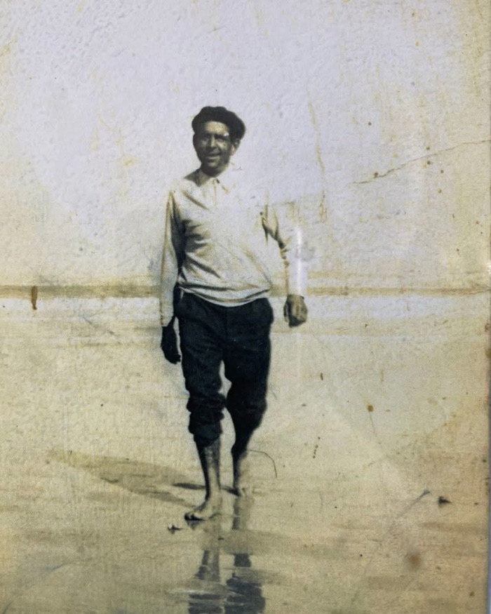 Acácio Francisco Grandela standing barefoot on wet sand in a white shirt and rolled trousers, in a cracked and faded photograph.