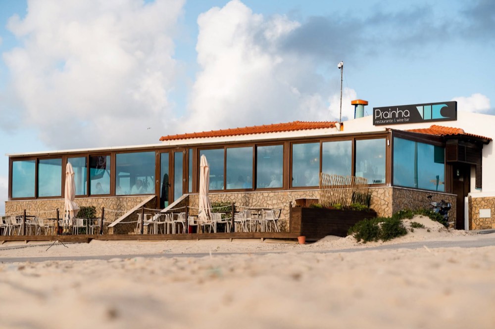 The Prainha restaurant, a low white building with a terracotta roof and glass frontage, sitting directly on the sand.