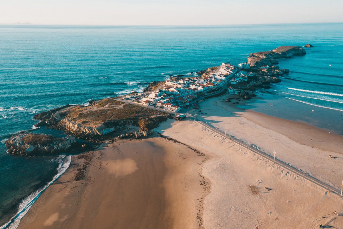Baleal from the air: the rocky headland and its white village joined to the mainland by a wide sand bar, with open ocean breaking on one side and calm water on the other.