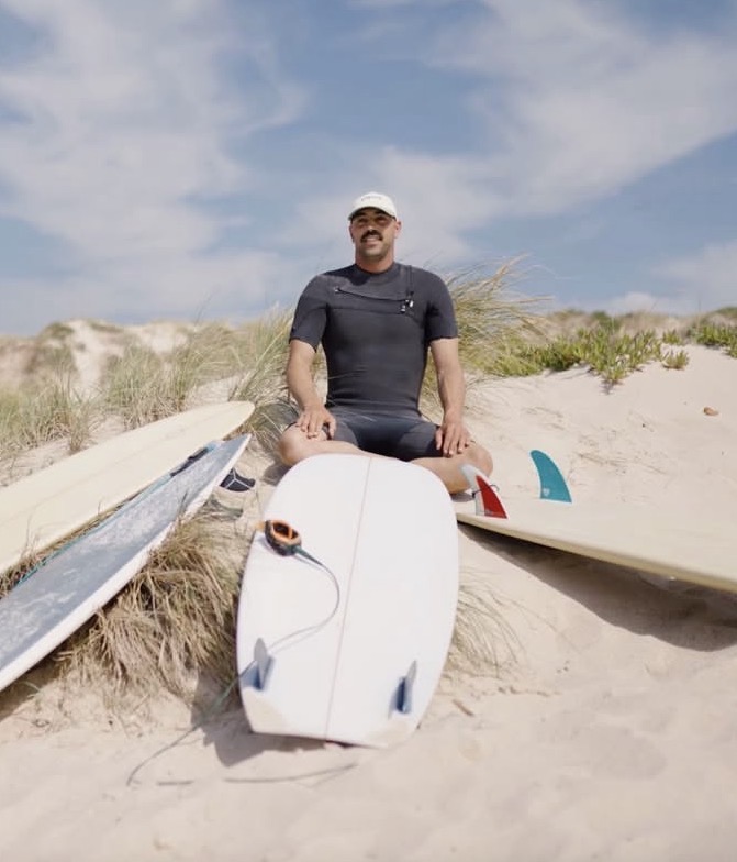 André sitting on the dunes in a wetsuit with three boards laid out beside him.