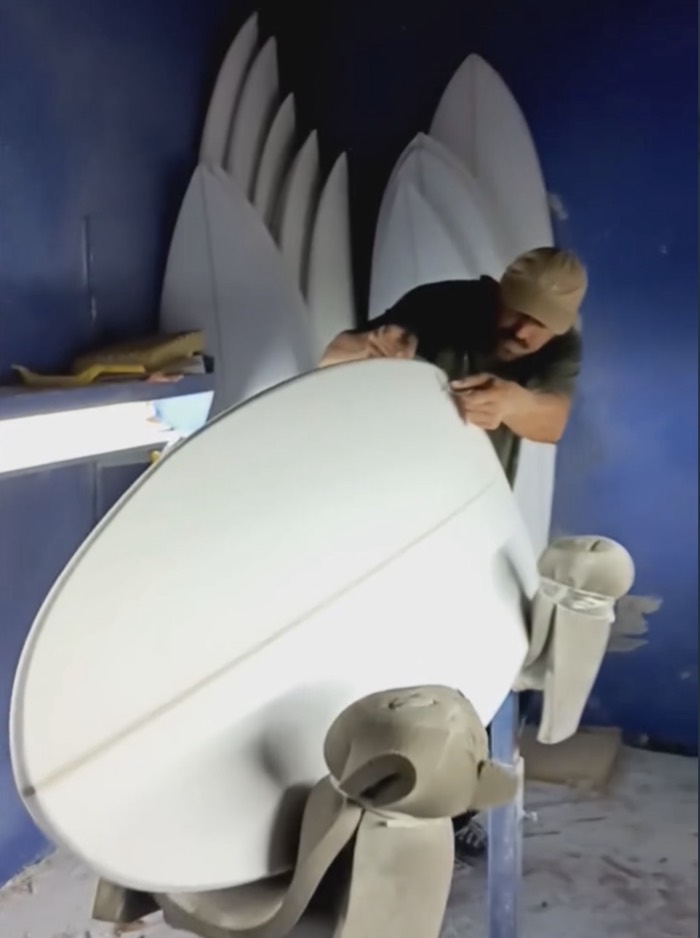 André shaping a surfboard blank in a shaping bay, more blanks stacked behind him.
