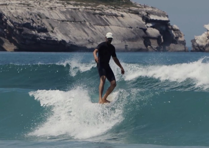 André walking the deck of a longboard on a clean blue wave, the Baleal cliffs behind him.