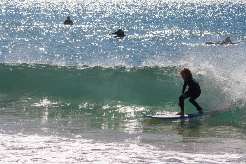 A young surfer riding a sunlit green wave as others paddle out behind