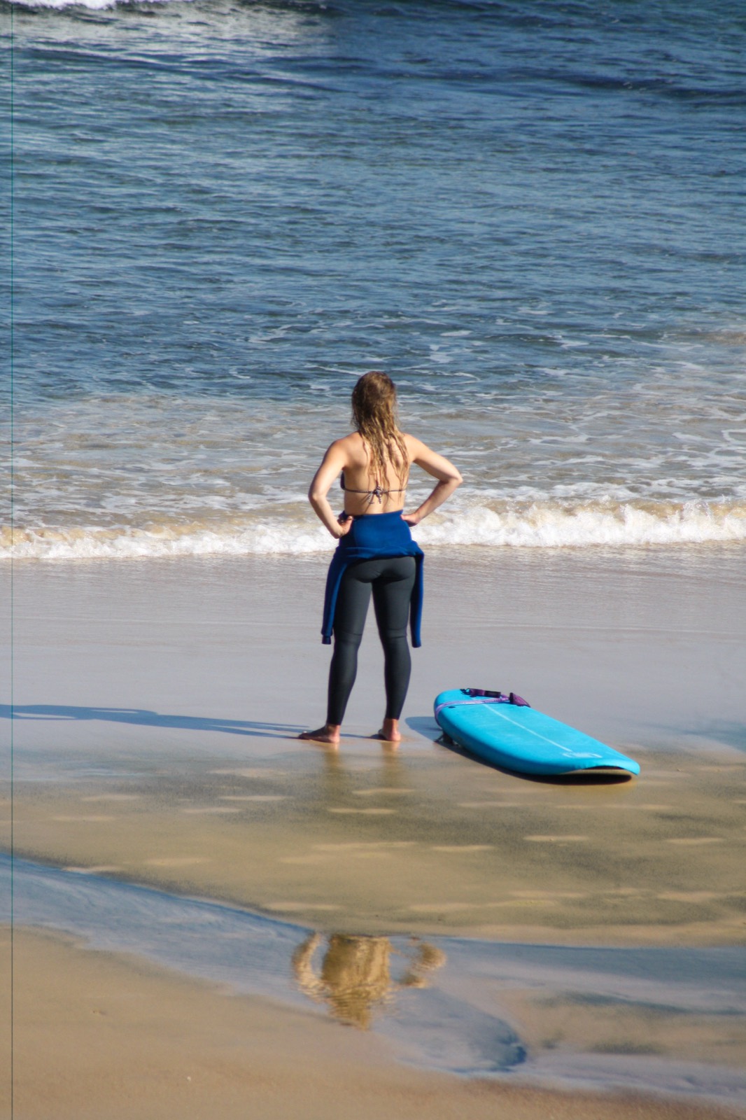 A surfer standing at the shoreline beside a blue softboard, looking at the sea