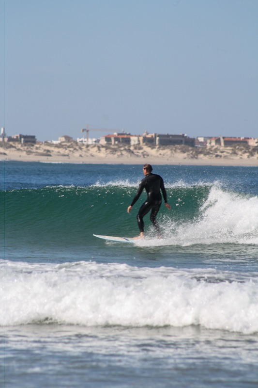 A surfer trimming along the wave face with the dunes and village behind